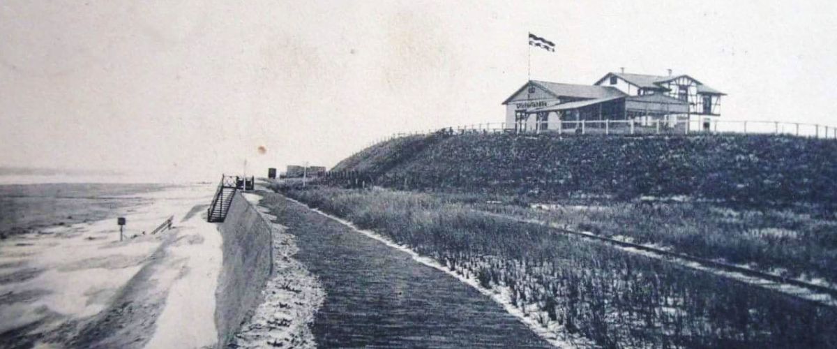 Ein historisches Strandcafé mit wehender Flagge thront über einem verschneiten Weg entlang der Küste.