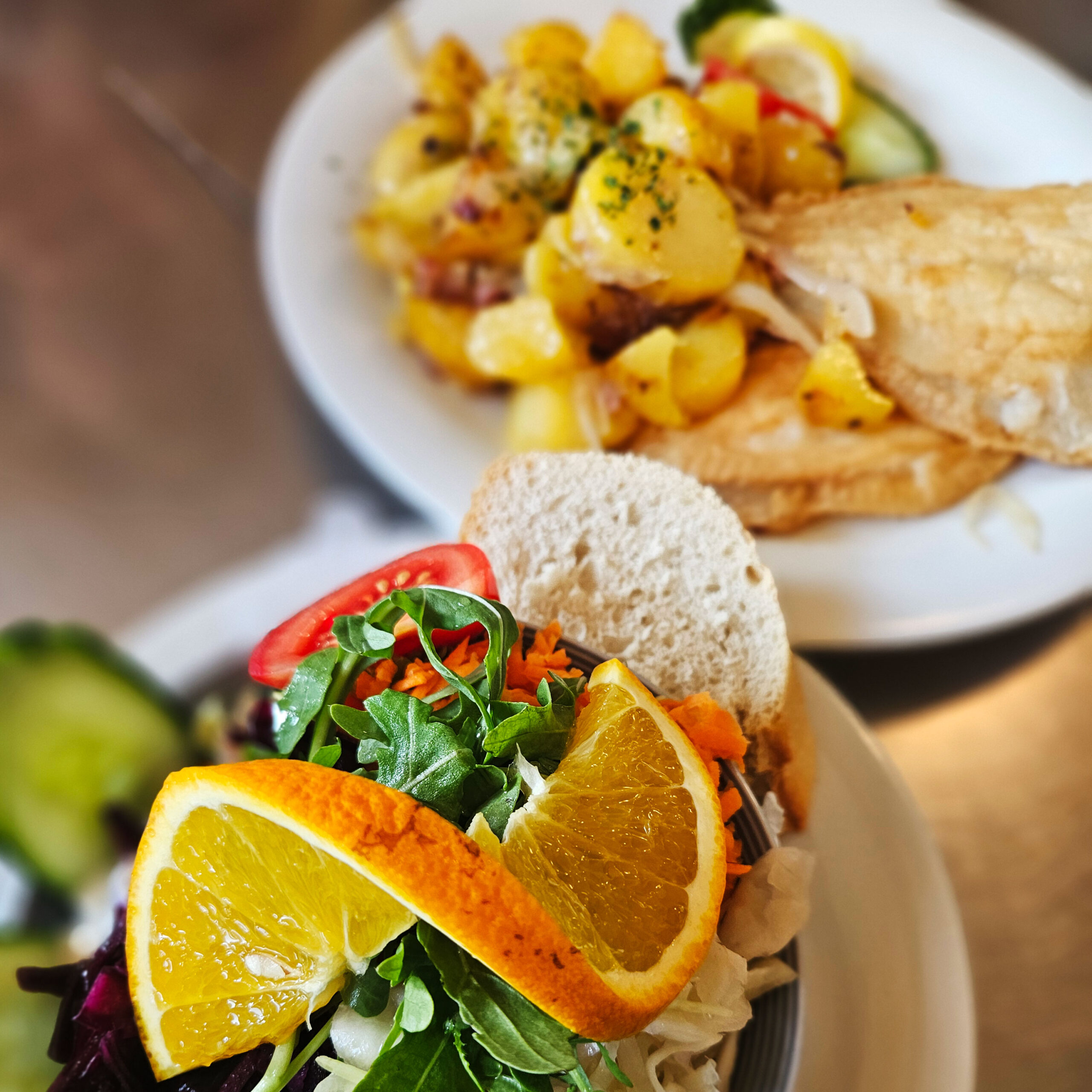 Ein frischer Salat mit Orangenscheiben und Brot wird neben einem Teller mit Bratkartoffeln und Fisch serviert.