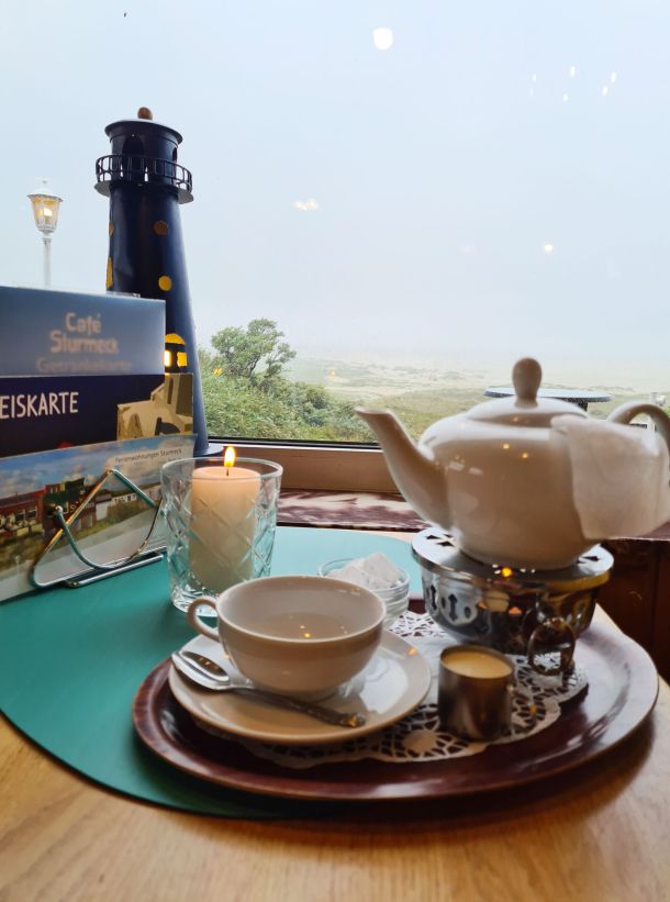 Eine gemütliche Teekanne steht auf einem Tisch im Strandcafé Sturmeck mit Blick auf einen Leuchtturm und die Dünenlandschaft.