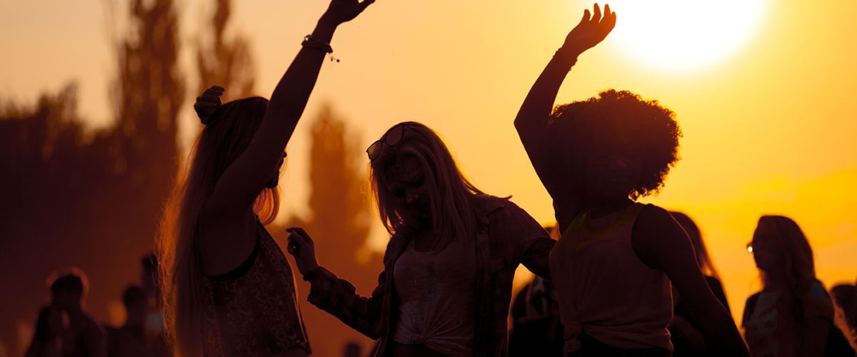 Drei Frauen tanzen fröhlich im Sonnenuntergang, umgeben von einer festlichen Atmosphäre und guter Laune.