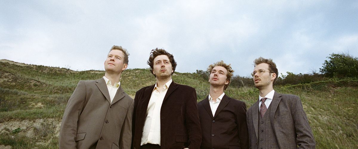 Vier Männer in Anzügen stehen auf einer grünen Wiese und blicken nachdenklich in den Himmel.