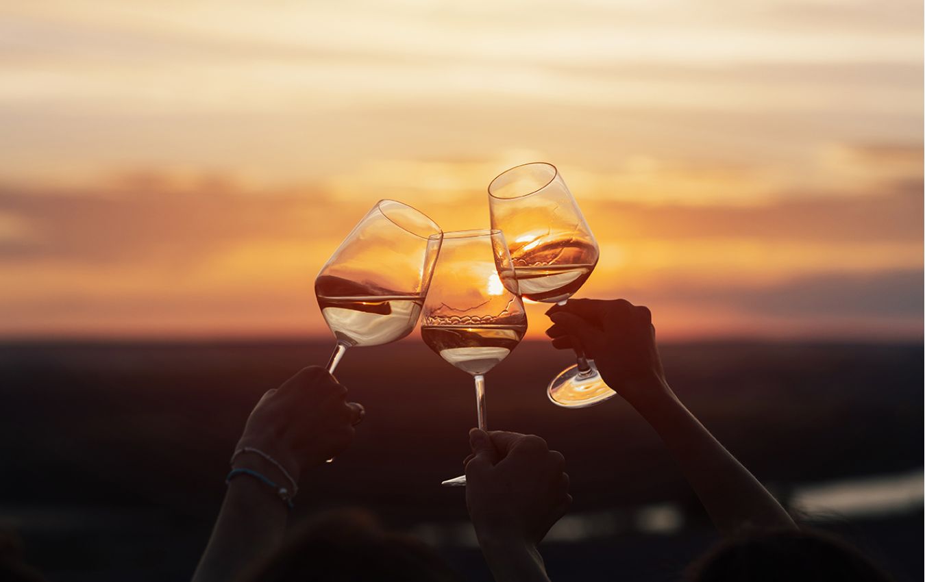 Drei Hände stoßen mit Weingläsern bei Sonnenuntergang an, passend zu einem festlichen Abend im Strandcafé.